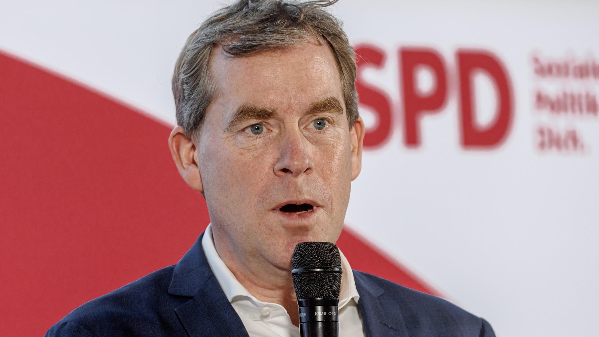 Das Bild zeigt den Kieler Bürgermeister Ulf Kämpfer, der vor einem SPD-Logo in ein Mikrofon spricht. 
