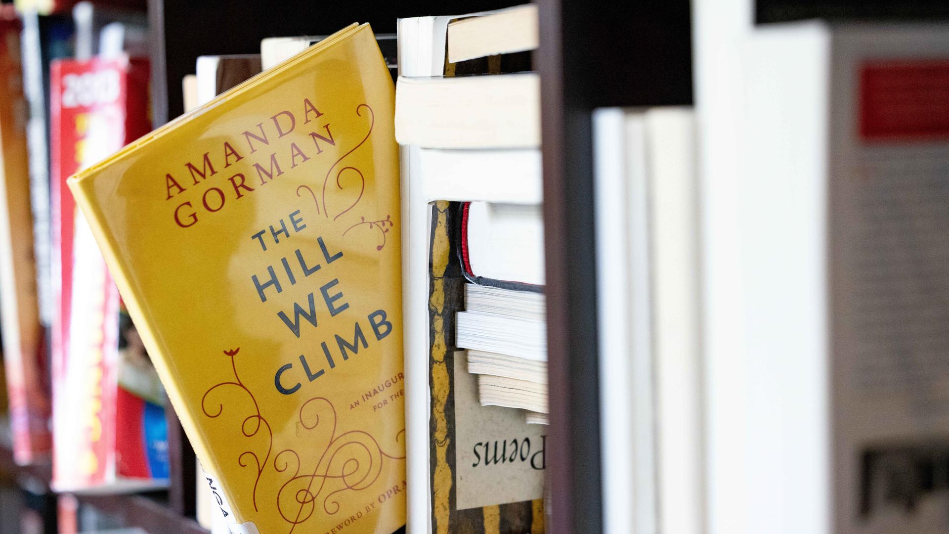 Ein gelbes Buch mit dem englischen  Titel "The hill we climb" scheint fast aus einem Bücherregal herauszufallen