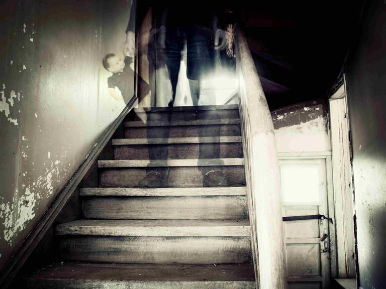 Eine Treppe in einem heruntergekommenen Haus, auf einer der Stufen steht die verschwommene, gespenstische Gestalt eines Mannes.