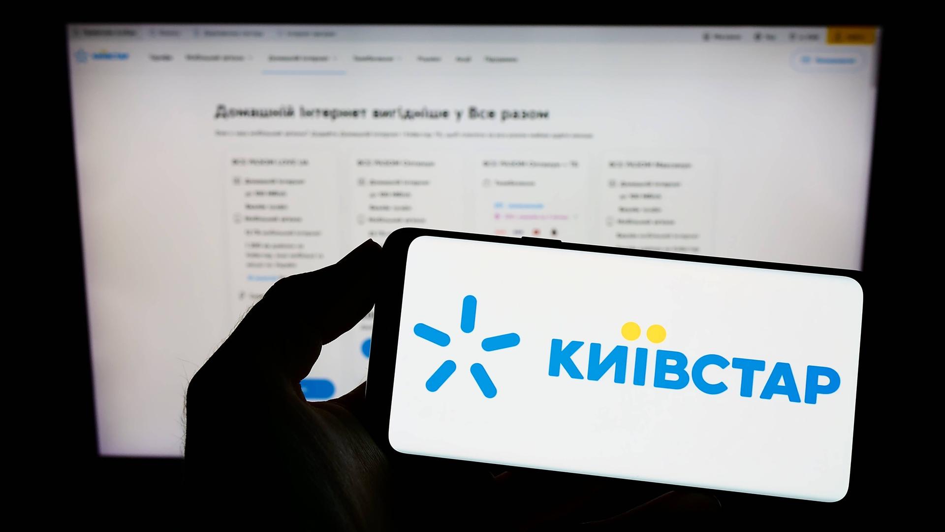 Eine Person hält ein Smartphone des ukrainischen Kommunikationsunternehmen Kyivstar in einer Hand. Dahinter verschwommen eine Internetseite.