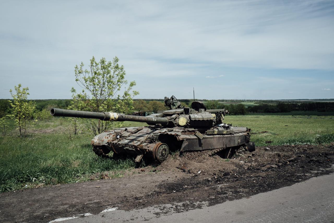 Der zerstörte Panzer steht an einem Straßenrand vor einem grünen Feld. 