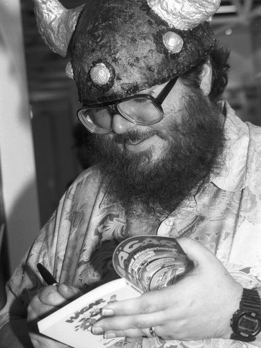 Schwarz-Weiß-Foto von Chris Browne, der einen Wikingerhelm trägt und einen Comicband von Hägar dem Schrecklichen signiert.