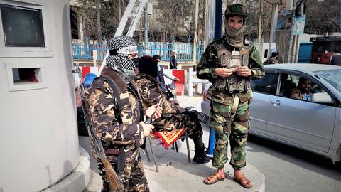 Zwei Taliban in grüner und brauner Tarnuniform stehen bewaffnet an einem Checkpoint in Kabul. Im Hintergrund sitzt ein weiterer Taliban auf einem Stuhl und schaut zu einem Autofahrer.