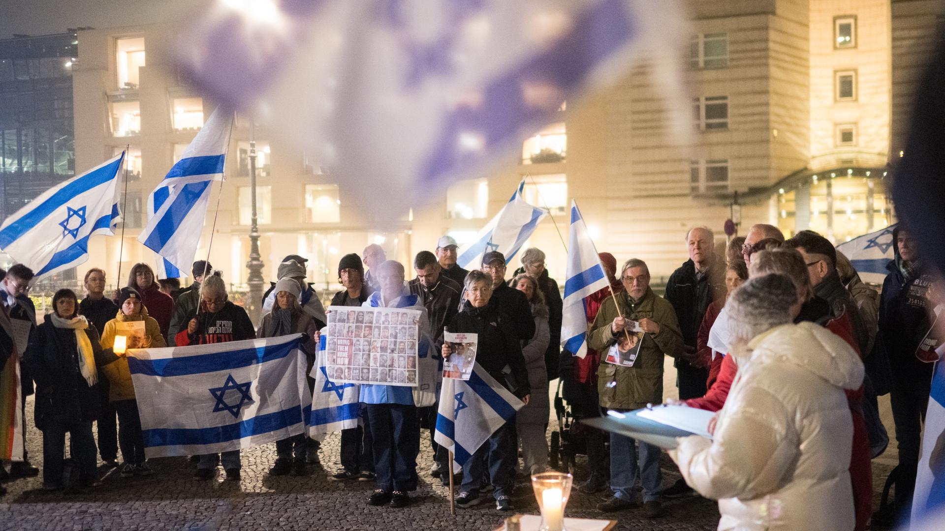 Menschen stehen in einer Gruppe zusammen, einige halten Israel-Flaggen in die Höhe - eine Frau am rechten Bildrand spricht, vor ihr steht eine Kerze.