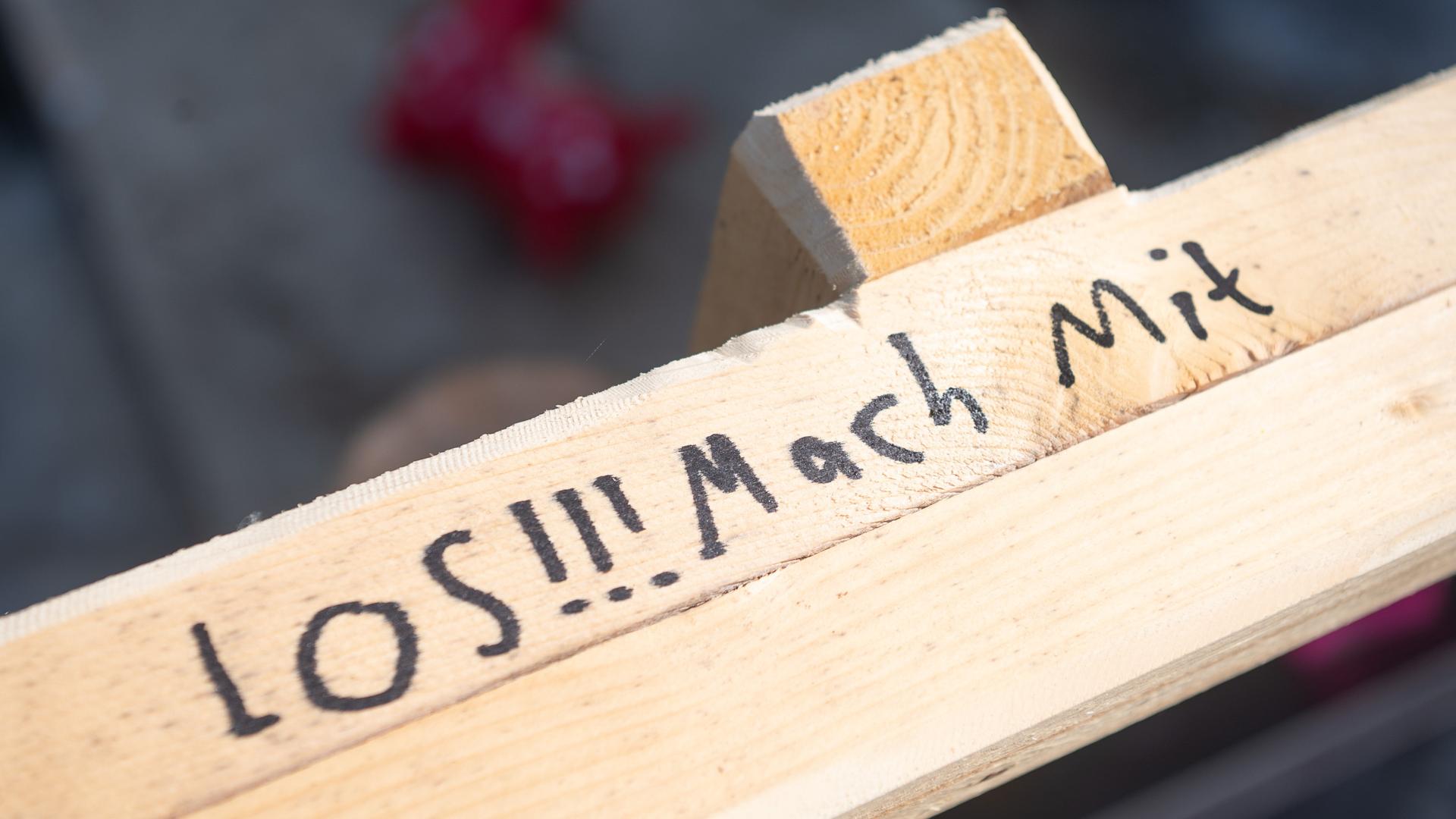 Auf einem Holbalken steht mit Edding "LOS!!! Mach Mit" geschrieben.