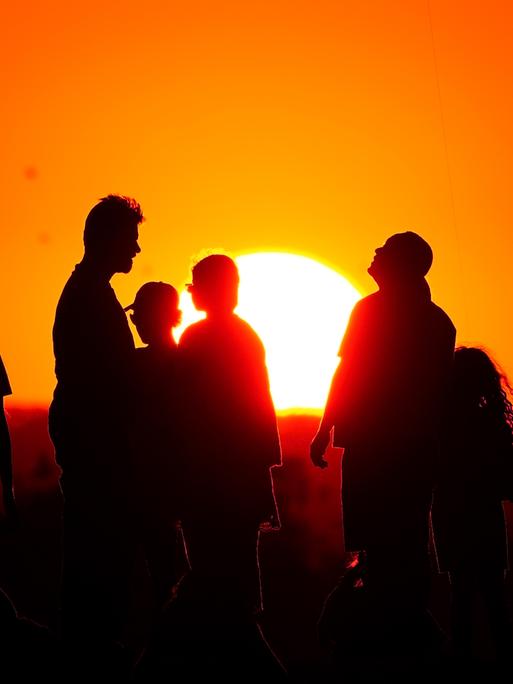 Eine Gruppe von Menschen steht vor der untergehenden Sonne am Horizont, man sieht nur ihre Schatten.