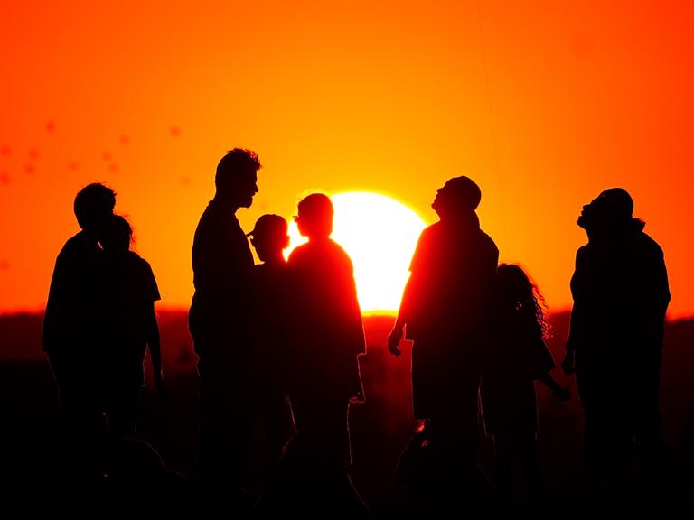 Eine Gruppe von Menschen steht vor der untergehenden Sonne am Horizont, man sieht nur ihre Schatten.