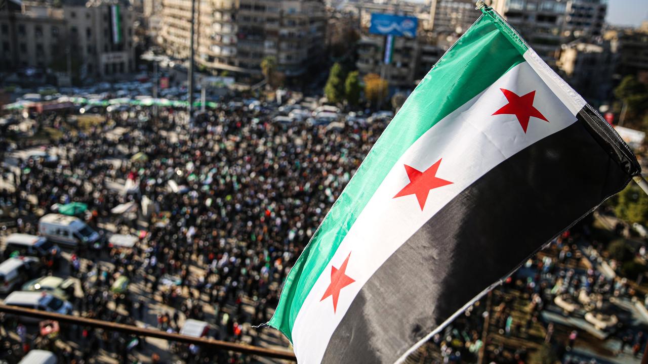 Menschen haben sich auf einem Platz in der Stadt Aleppo versammelt, um den Sturz des Assad-Regimes zu feiern. Über ihnen weht die neue syrische Flagge mit drei roten Sternen.  