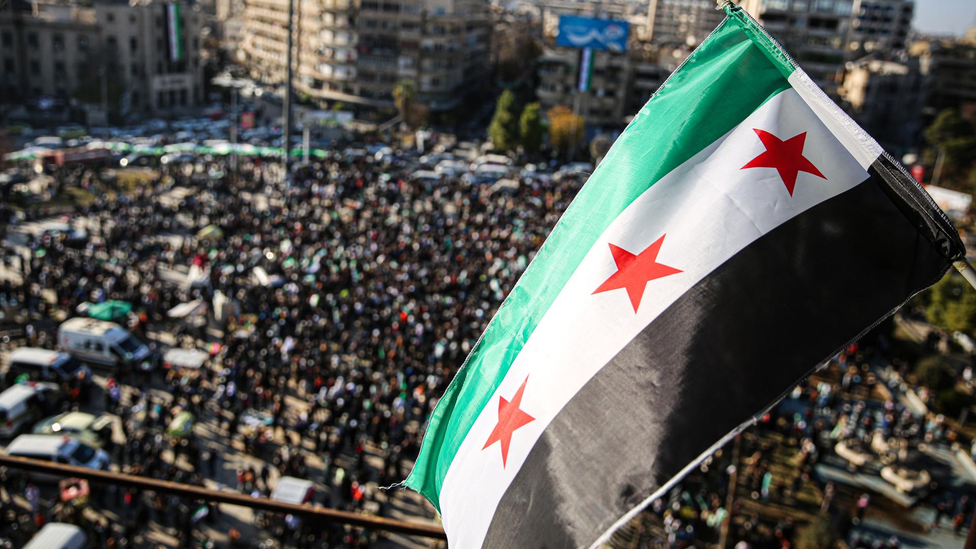 Menschen haben sich auf einem Platz in der Stadt Aleppo versammelt, um den Sturz des Assad-Regimes zu feiern. Über ihnen weht die neue syrische Flagge mit drei roten Sternen. 