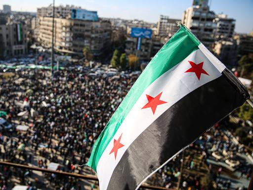 Menschen haben sich auf einem Platz in der Stadt Aleppo versammelt, um den Sturz des Assad-Regimes zu feiern. Über ihnen weht die neue syrische Flagge mit drei roten Sternen.  