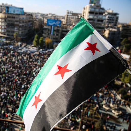 Menschen haben sich auf einem Platz in der Stadt Aleppo versammelt, um den Sturz des Assad-Regimes zu feiern. Über ihnen weht die neue syrische Flagge mit drei roten Sternen. 