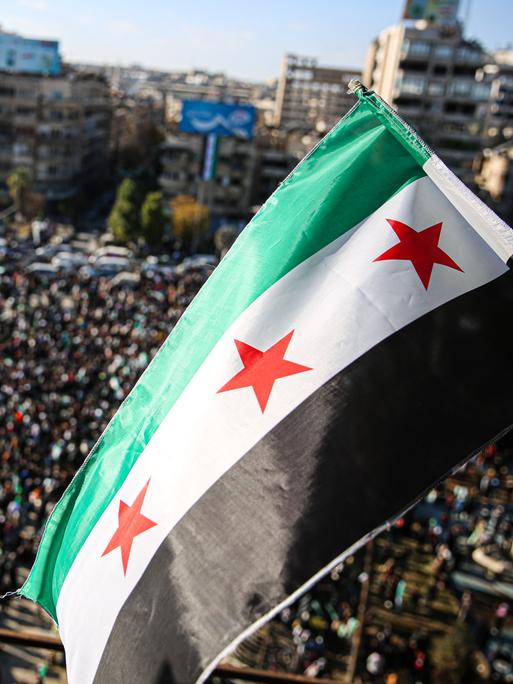 Menschen haben sich auf einem Platz in der Stadt Aleppo versammelt, um den Sturz des Assad-Regimes zu feiern. Über ihnen weht die neue syrische Flagge mit drei roten Sternen.  