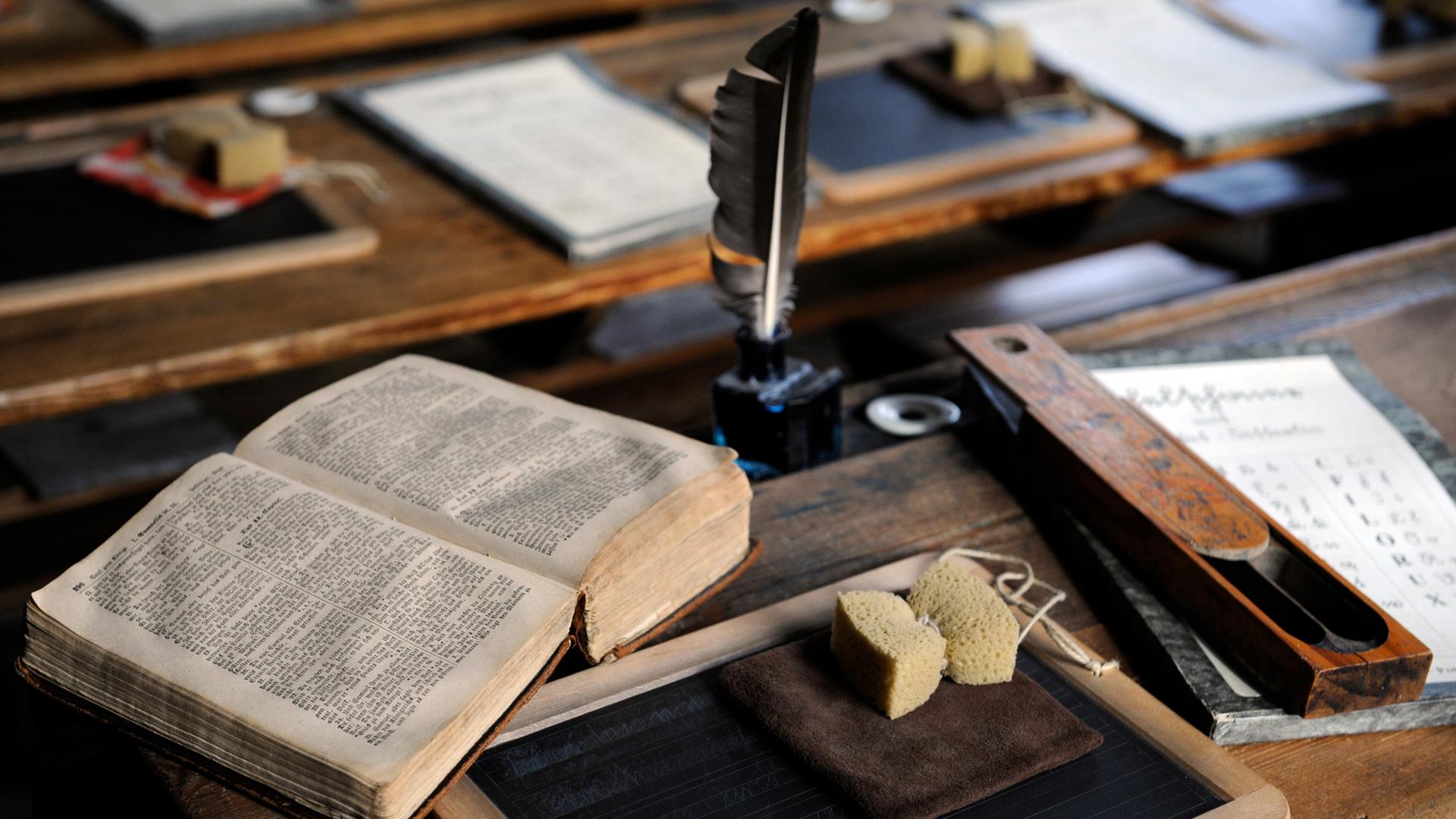 Historische Schulsachen wie eine Schreibfeder, Schiefertafel, Schwamm und Buch, liegen auf einem Schreibpult