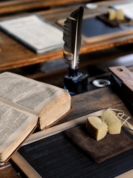 Historische Schulsachen wie eine Schreibfeder, Schiefertafel, Schwamm und Buch, liegen auf einem Schreibpult
