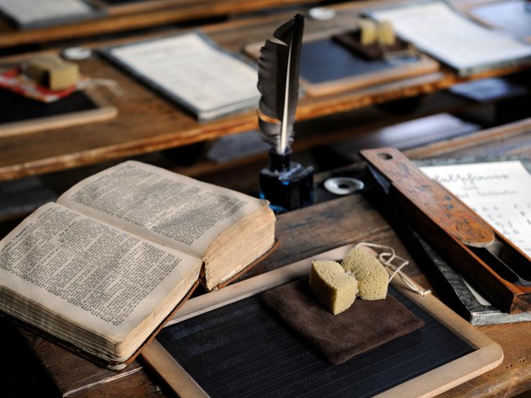 Historische Schulsachen wie eine Schreibfeder, Schiefertafel, Schwamm und Buch, liegen auf einem Schreibpult