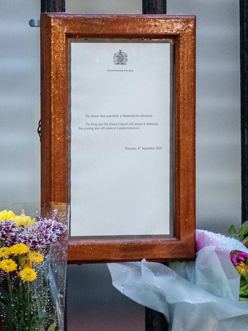 Ein Blatt Papier mit der Todesanzeige von Königin Elisabeth hängt in einem braunen Rahmen an einem Zaun, daneben stehen Blumen.