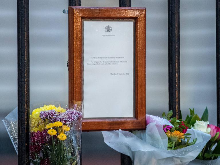 Ein Blatt Papier mit der Todesanzeige von Königin Elisabeth hängt in einem braunen Rahmen an einem Zaun, daneben stehen Blumen.