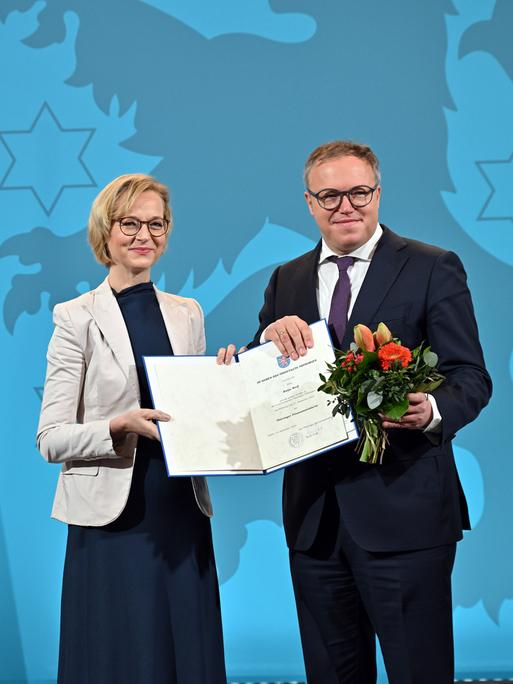 Thüringens Finanzministerin Katja Wolf (BSW) und Ministerpräsident Mario Voigt (CDU) mit Wolfs Ernennungsurkunde und einem Blumenstrauß. Links sieht man die weiße-rote Landesfahne mit dem Löwen im blauem Wappen.