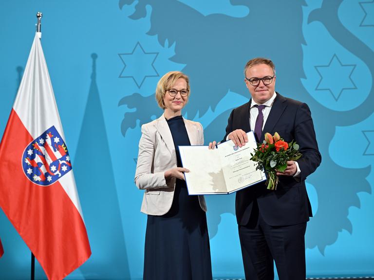 Thüringens Finanzministerin Katja Wolf (BSW) und Ministerpräsident Mario Voigt (CDU) mit Wolfs Ernennungsurkunde und einem Blumenstrauß. Links sieht man die weiße-rote Landesfahne mit dem Löwen im blauem Wappen.