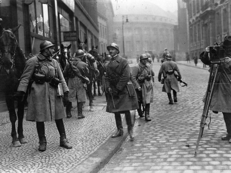 Französische Soldaten in Essen bei der Besetzung des Ruhrgebiets durch französische Truppen. Links hält ein Soldat ein Pferd, andere Soldaten gehen in der Straße umher, ganz rechts steht ein Fotograf mit seiner aufgebauten Kamera.