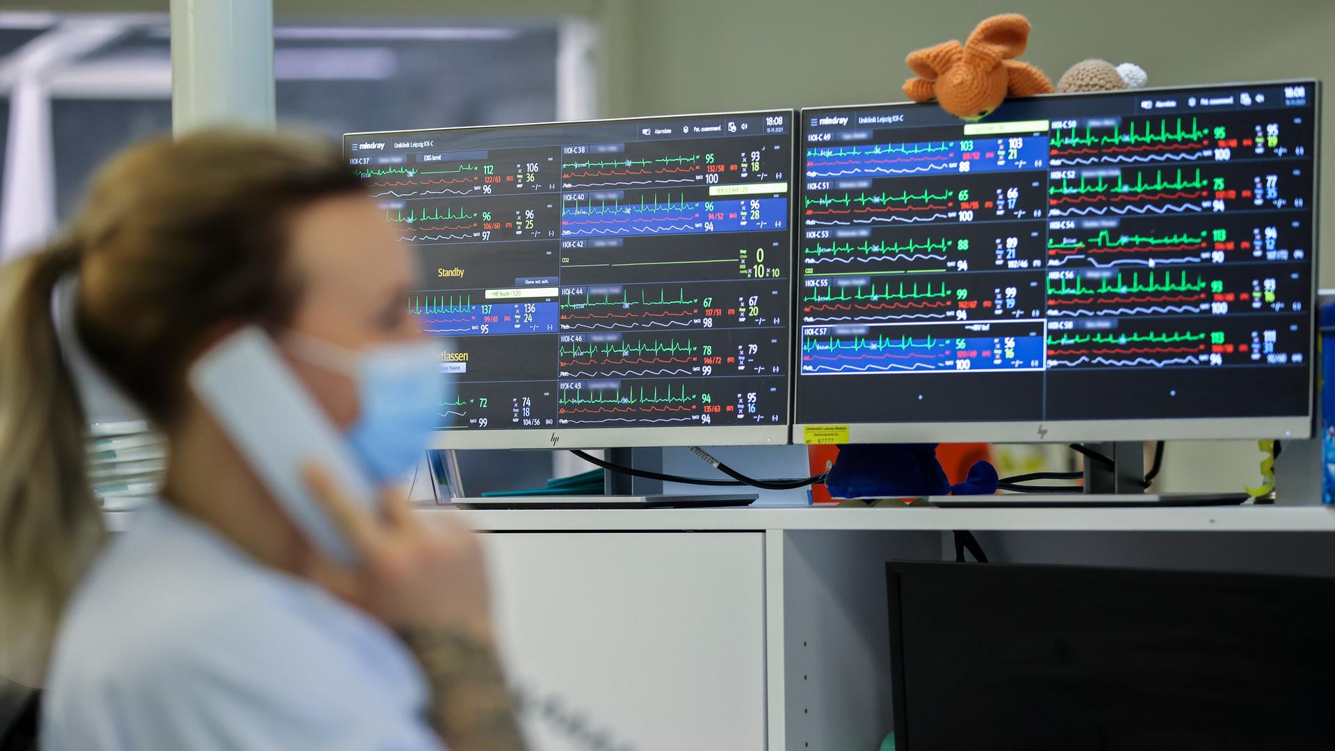 Unscharf im Vordergrund Intenstivpflegerin am Telefon, scharf im Hintergrund Monitore mit mehreren EKGs von Intensivpatientinnen und -patienten Unscharf im Vordergrund Intenstivpflegerin am Telefon, scharf im Hintergrund Monitore mit mehreren EKGs von Intensivpatientinnen und -patienten