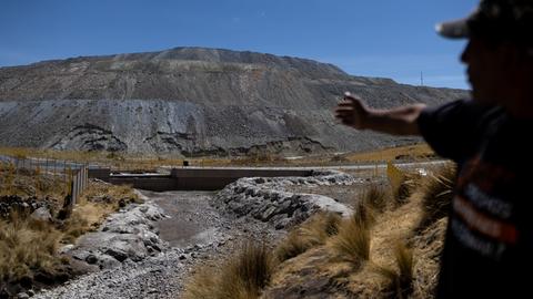 Vidal Merma steht nahe einer Kupfermine in Espinar. Nur sein Schatten ist erkennbar. Er zeigt auf Abfälle. Im Hintergrund sind Zäune und ein grauer Berg aus Steinen und Sand zu sehen.