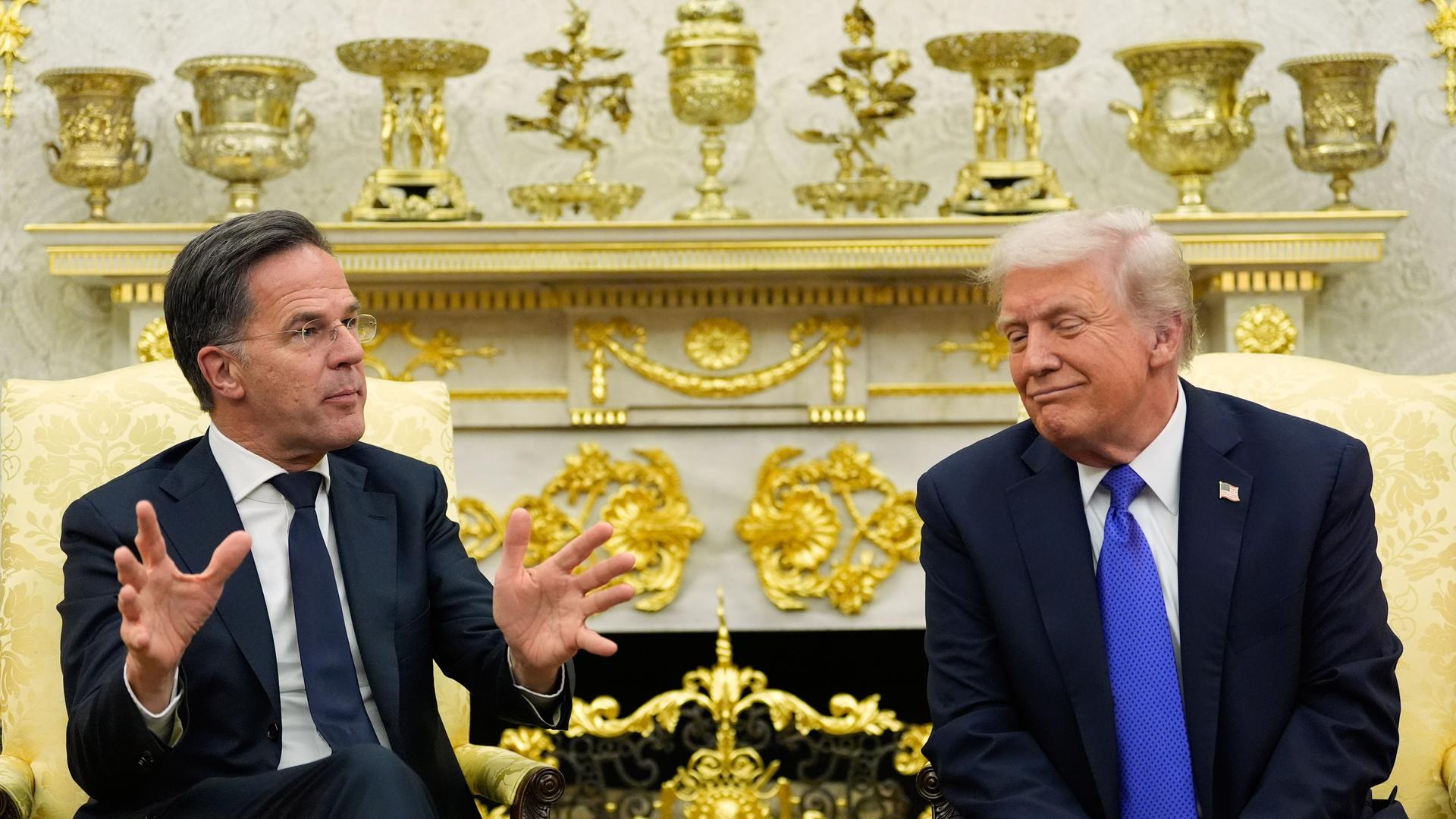 NATO-Generalsekretär Mark Rutte und US-Präsident Donald Trump sitzen nebeneinander. Rutte gestikuliert. Trump hat seinen Kopf zu ihm hin gewandt. Im Hintergrund stehen auf einem Kaminsims unter anderem goldfarbene Pokale.