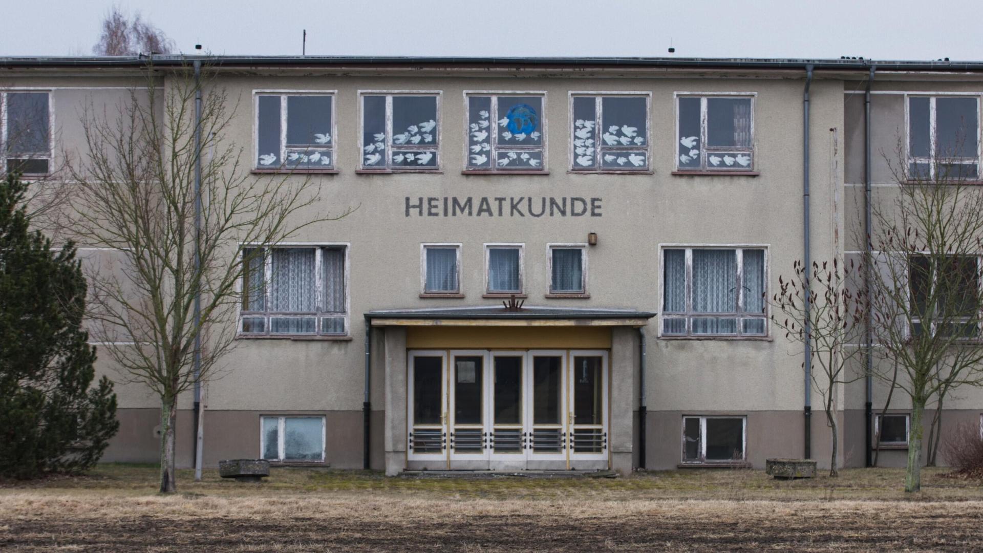 Ein großes, graues Gebäude auf einem winterlichen Feld. Es handelt sich um die Polytechnische Oberschule Bärenklau. Das Foto ist die Titeleinstellung des Films und zeigt die Front mit Eingangstür. Darüber steht der Schriftzug "Heimatkunde".