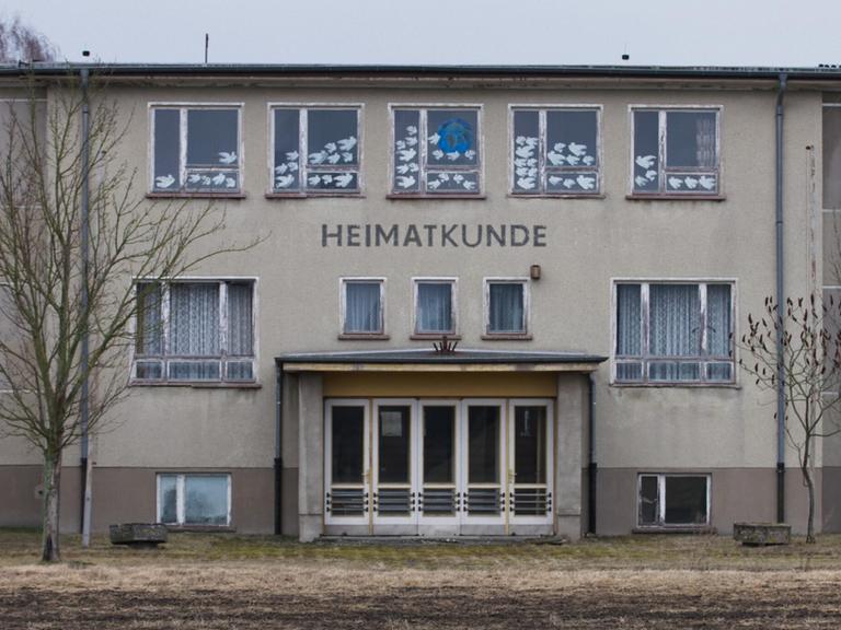 Ein großes, graues Gebäude auf einem winterlichen Feld. Es handelt sich um die Polytechnische Oberschule Bärenklau. Das Foto ist die Titeleinstellung des Films und zeigt die Front mit Eingangstür. Darüber steht der Schriftzug "Heimatkunde".