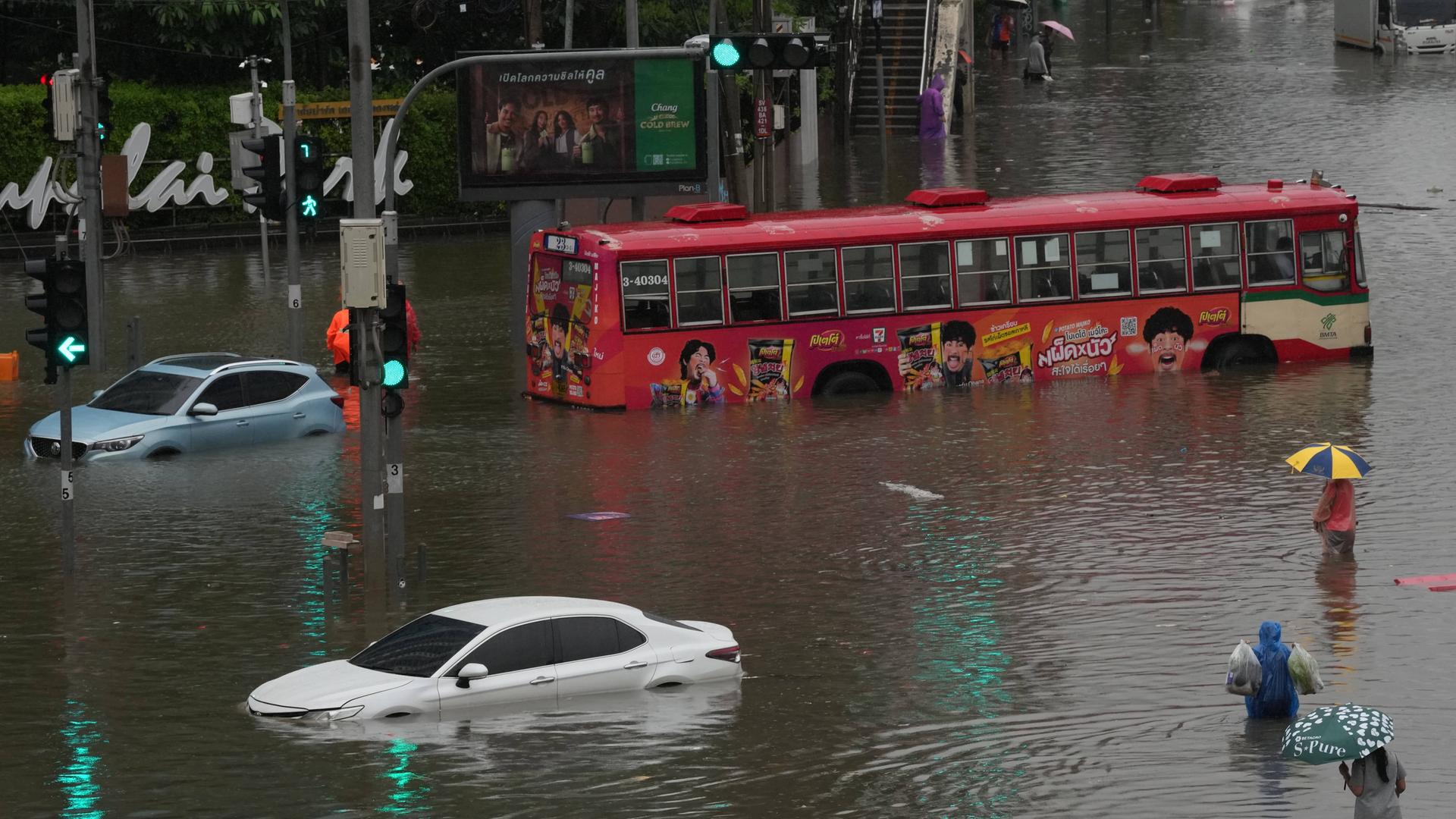 Eine Straße in Bangkok steht unter Wasser. Ein Bus und mehrere Autos sind halb überschwemmt. Fußgänger waten durch das Wasser. 