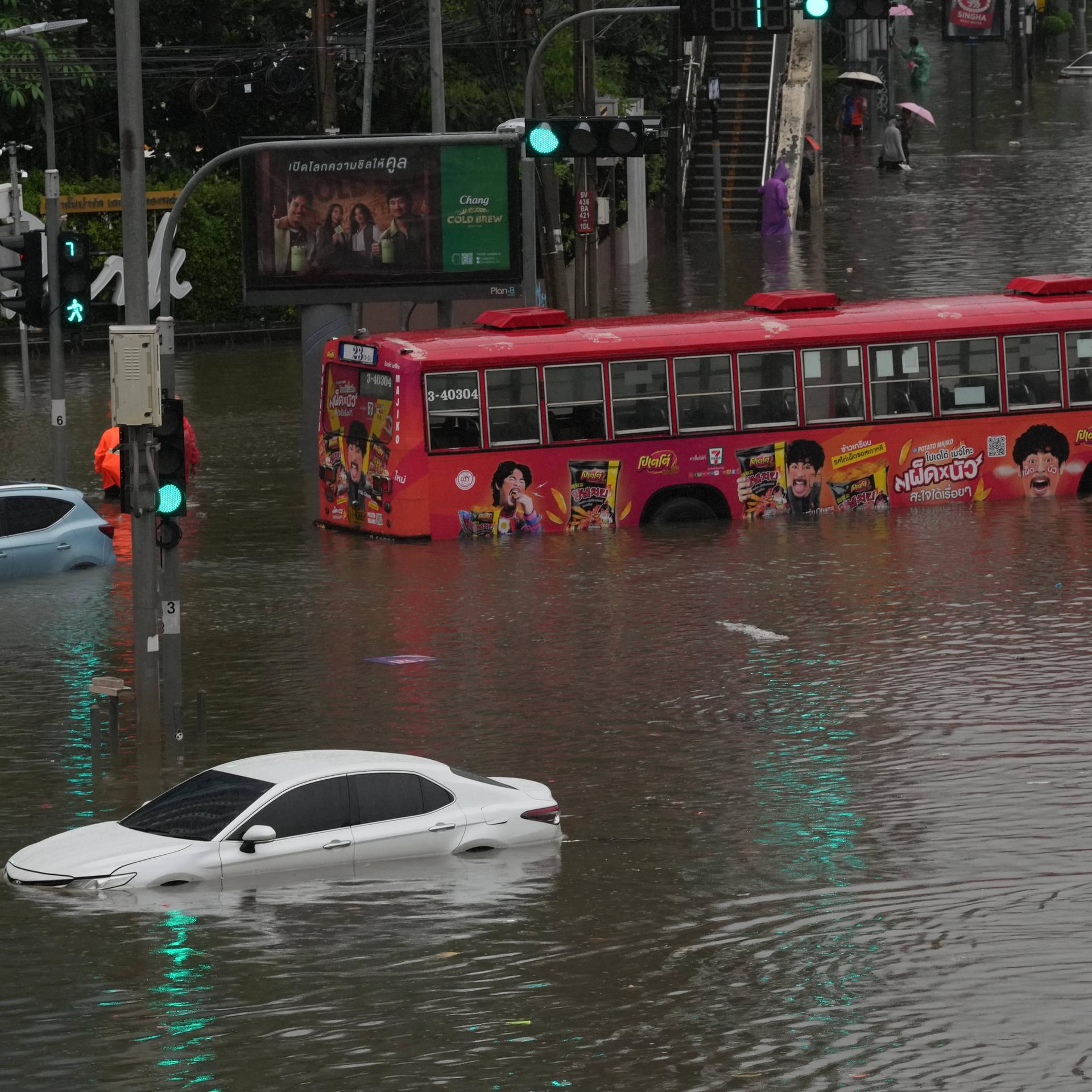 Eine Straße in Bangkok steht unter Wasser. Ein Bus und mehrere Autos sind halb überschwemmt. Fußgänger waten durch das Wasser. 