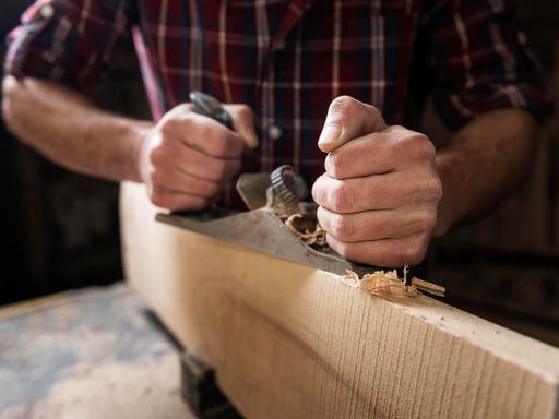 Ein Mann mit kariertem Hemd arbeitet mit Holz. Seine Hand hält einen Hobel, es fallen Späne. 