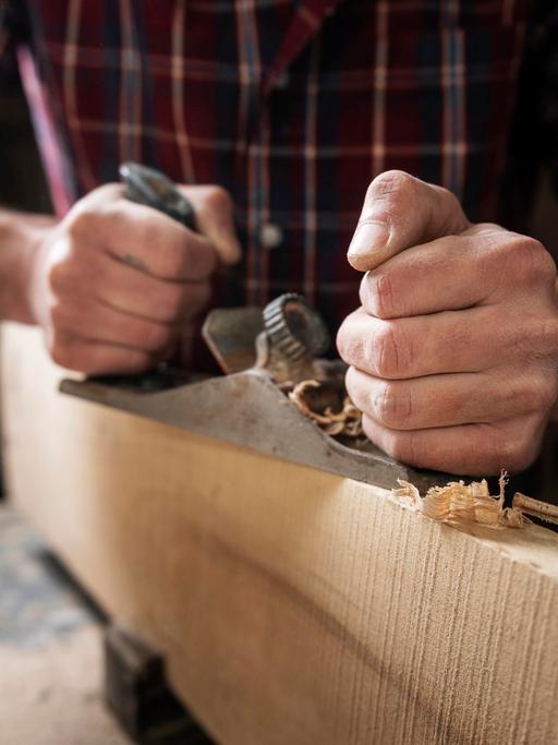 Ein Mann mit kariertem Hemd arbeitet mit Holz. Seine Hand hält einen Hobel, es fallen Späne. 