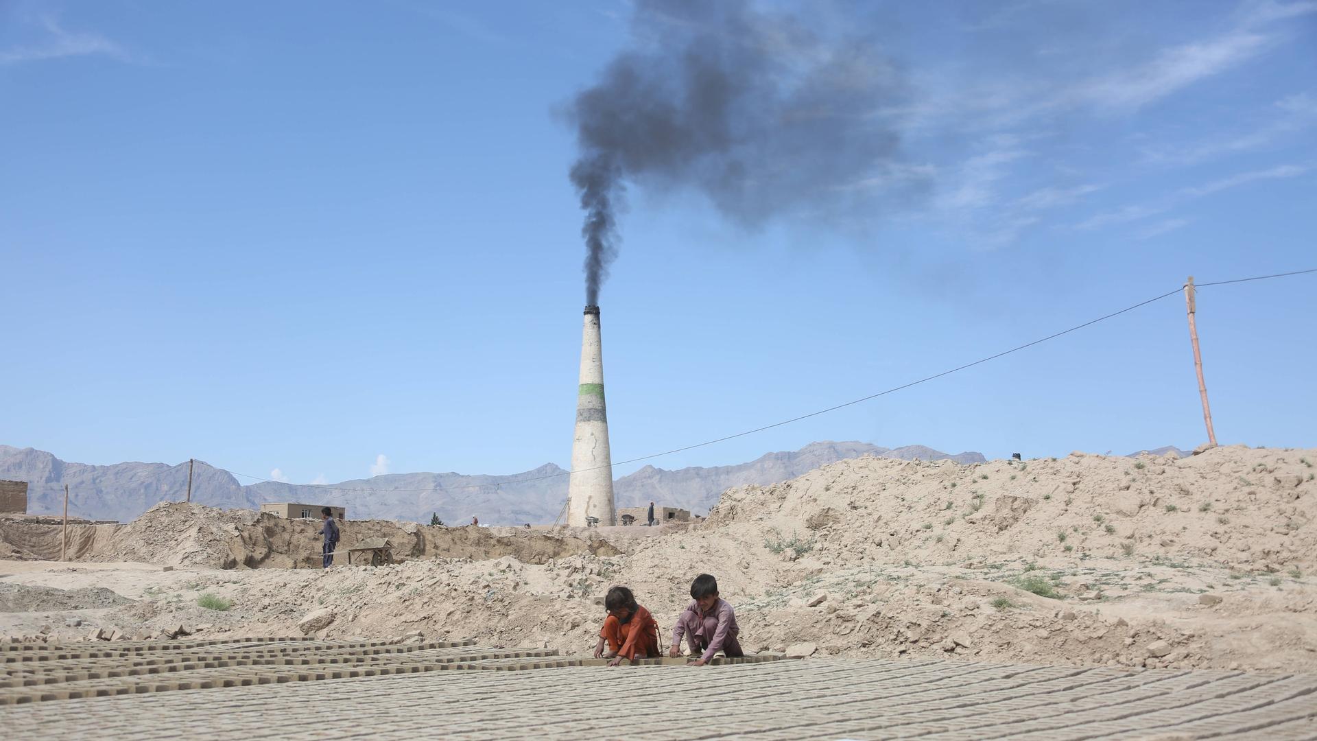 Zwei Kinder sitzen auf dem Boden und arbeiten mit ihren Händen. Im Hintergrund ist ein Schornstein. Es ist eine Ziegelei in Afghanistan.