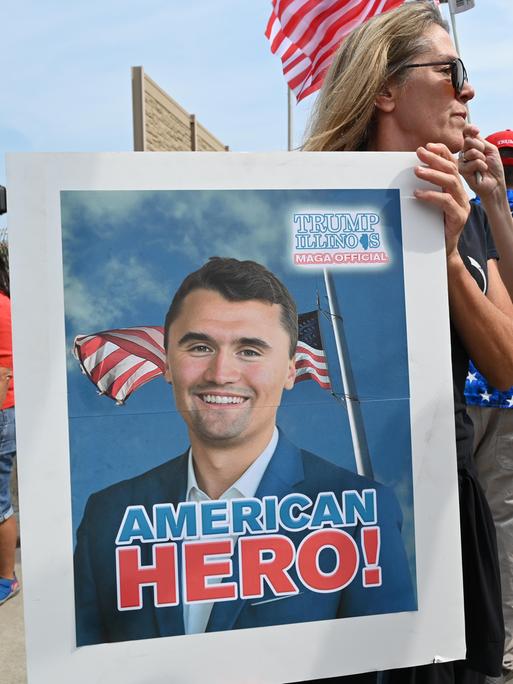 Eine Frau trägt ein Plakat mit dem Bild von Charlie Kirk, auf dem eine amerikanische Flagge zu sehen ist und die Worte "American Hero!" 