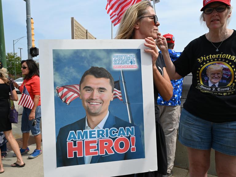Eine Frau trägt ein Plakat mit dem Bild von Charlie Kirk, auf dem eine amerikanische Flagge zu sehen ist und die Worte "American Hero!" 