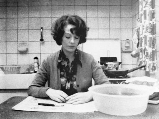 Auf dem Schwarz-Weiß-Bild sitzt eine Frau am Küchentisch. Vor ihr eine Schüssel mit Wasser und ein Sparschäler. Sie blickt nach unten, sie wirkt in sich gekehrt. 