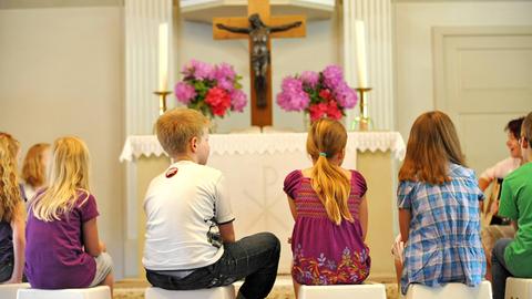 Mädchen und Jungen sitzen in einer Kirche vor dem Altar auf kleinen Hockern und blicken auf das Kreuz. Man sieht sie von hinten. 
