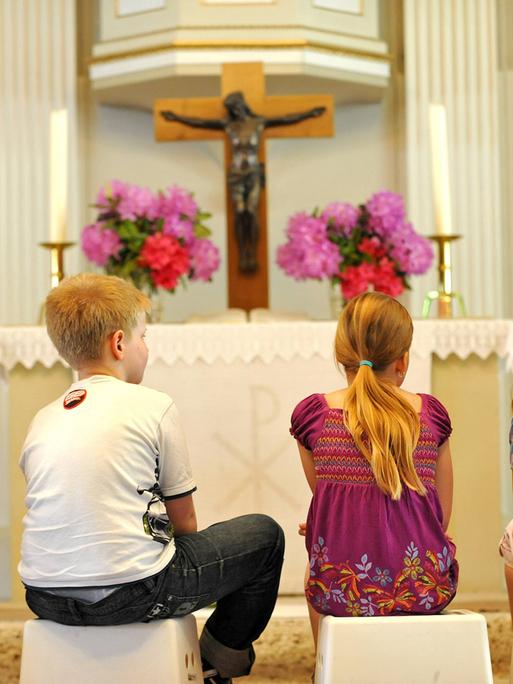 Mädchen und Jungen sitzen in einer Kirche vor dem Altar auf kleinen Hockern und blicken auf das Kreuz. Man sieht sie von hinten. 