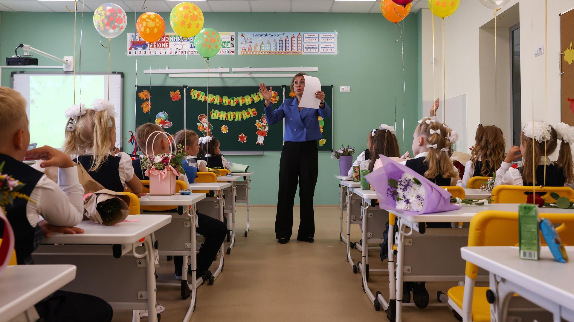 Eine Lehrerin steht am ersten Schultag in einem Klassenzimmer der ersten Klasse und erklärt den Schülerinnen und Schülern etwas. Die Kinder haben Luftballons an ihre Bänke gebunden. Eine Lehrerin steht am ersten Schultag in einem Klassenzimmer der ersten Klasse und erklärt den Schülerinnen und Schülern etwas. Die Kinder haben Luftballons an ihre Bänke gebunden.