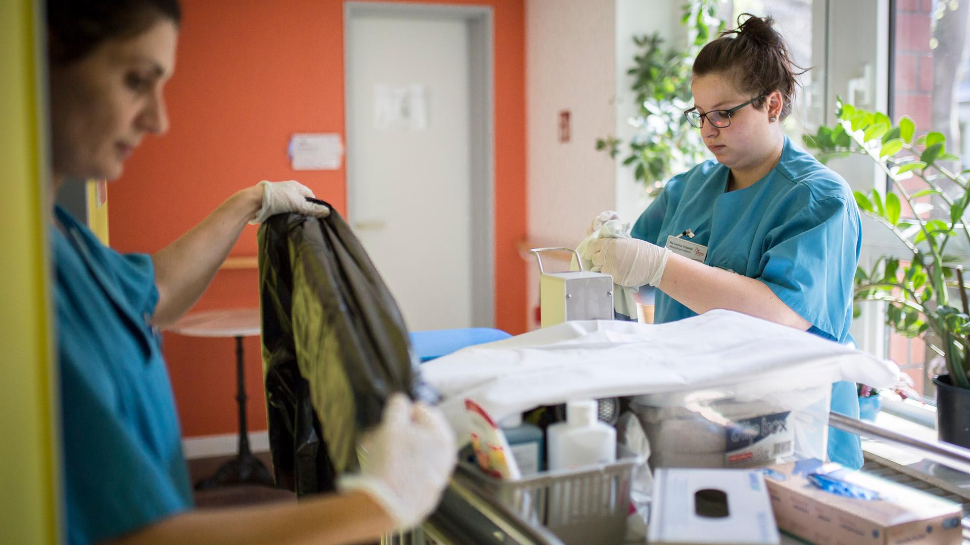 Zwei Pflegerinnen stehen an einem Wäsche-Wagen in einem Altenheim. Sie tragen hellblaue Kittel. 