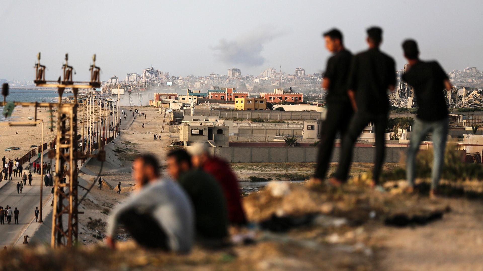 Vom Gazastreifen steigt Rauch auf, der aus der Ferne zu sehen ist. Im Vordergrund sitzen junge Männer und schauen auf die Stadt.