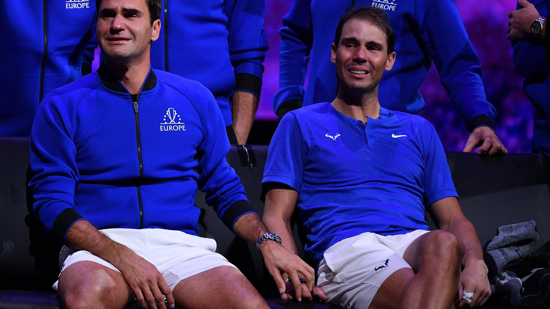 Federer und Nadal sitzen weinend in blauen Oberteilen auf eine Bank und Federer legt seine Hand auf Nadals. 