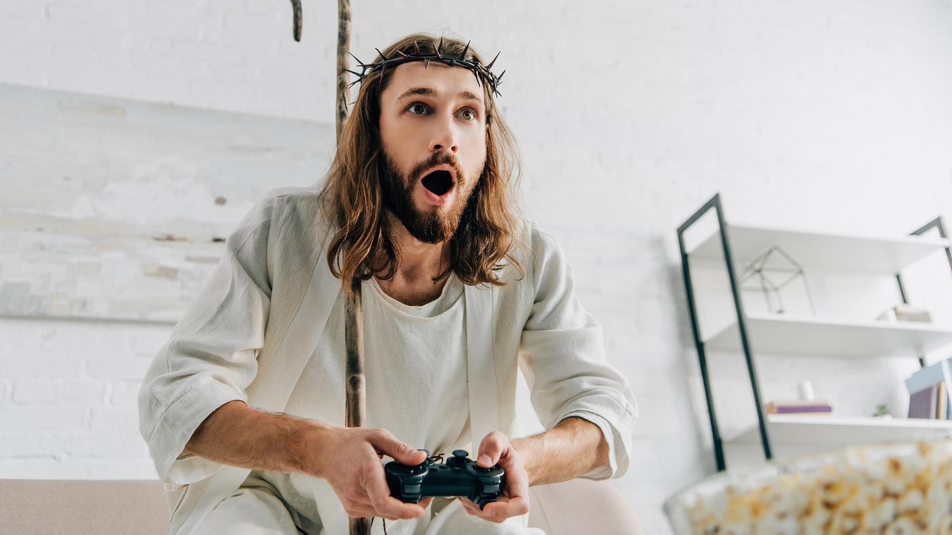 Ein Mann, der gekleidet ist wie eine Jesusfigur mit Dornenkrone und weißer Bekleidung, sitzt begeistert vor einem Videospiel.