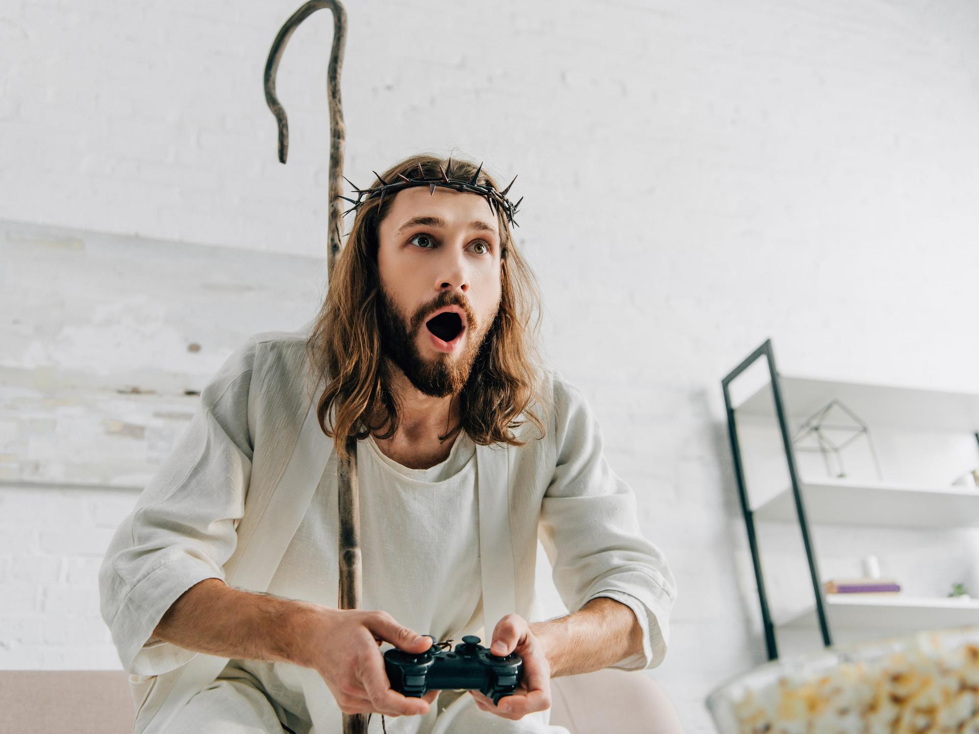 Ein Mann, der gekleidet ist wie eine Jesusfigur mit Dornenkrone und weißer Bekleidung, sitzt begeistert vor einem Videospiel.