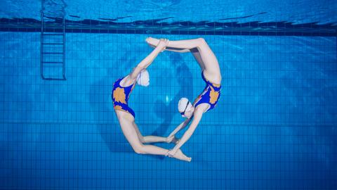 Zwei Synchronschwimmen bilden in einem Swimmingpool unter Wasser einen Kreis.