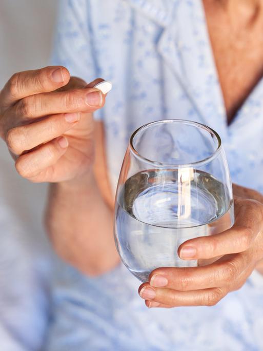 Eine ältere Frau hält eine Tablette und ein Wasserglas in der Hand, während sie im Schlafanzug auf einem Krankenhausbett sitzt.