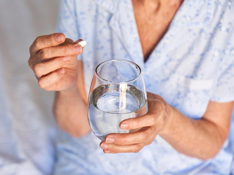 Eine ältere Frau hält eine Tablette und ein Wasserglas in der Hand, während sie im Schlafanzug auf einem Krankenhausbett sitzt.
