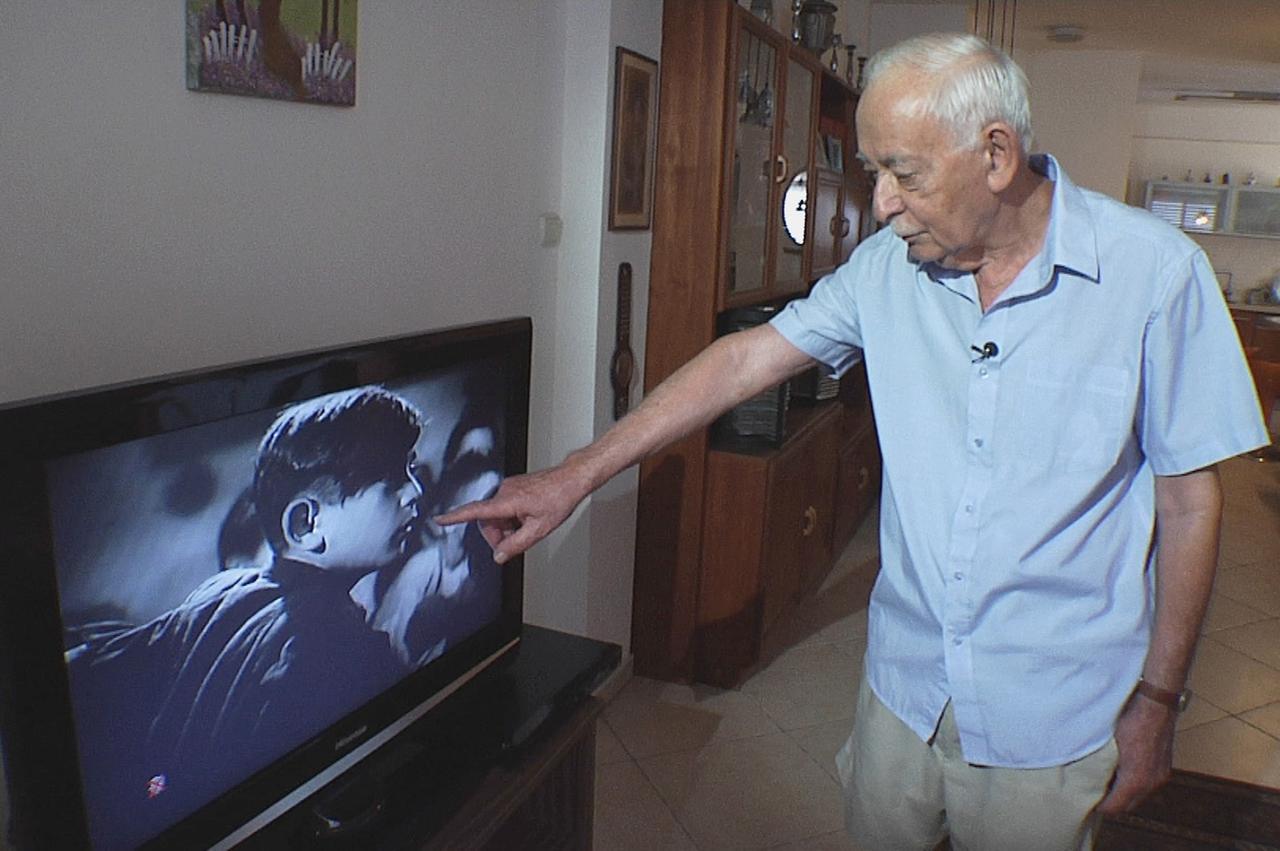 Ein alter, schlanker Mann mit weißen Haaren zeigt auf das Bild im Fernseher. Dort sind mehrere Kinder in Großaufnahme zu sehen.