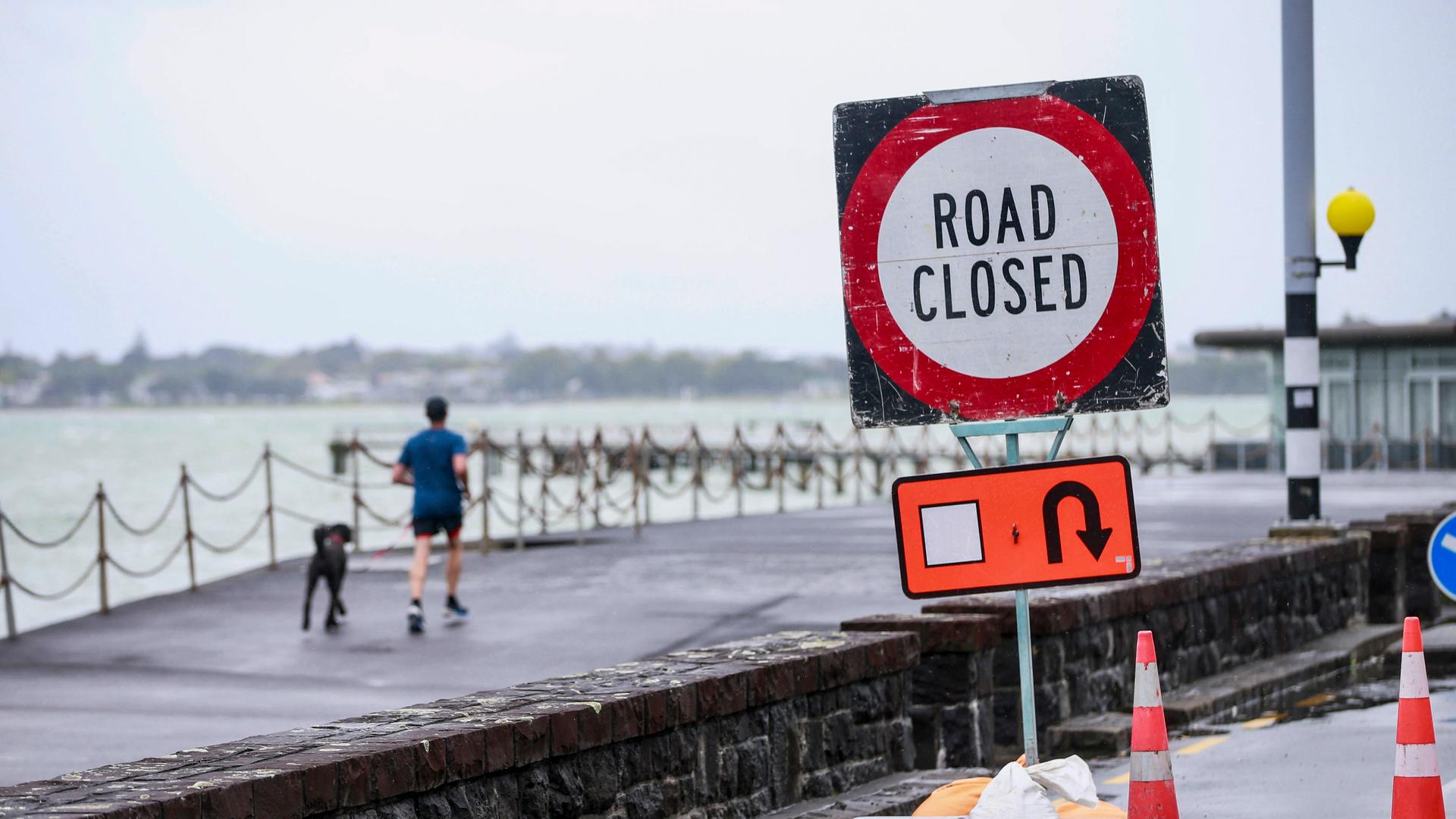 Man sieht ein Schild mit der Aufschrift "Straße gesperrt". Im Hintergrund ist ein Wellenbrecher, auf dem ein Mann joggt. Man sieht ein Schild mit der Aufschrift "Straße gesperrt". Im Hintergrund ist ein Wellenbrecher, auf dem ein Mann joggt.